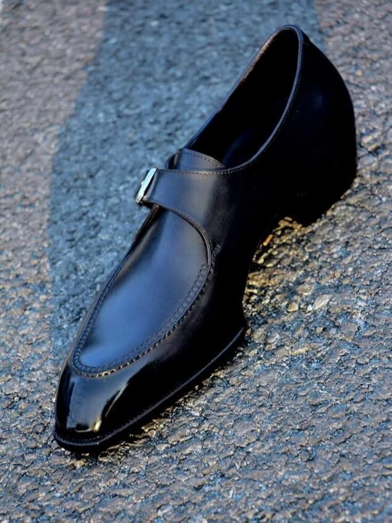 May include: A black leather dress shoe with a pointed toe and a strap with a silver buckle. The shoe has a glossy finish and is set against a textured surface.