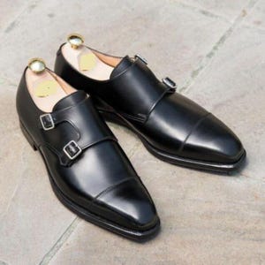 May include: A pair of black leather dress shoes with a double monk strap design. The shoes have a polished finish, a cap toe, and silver-colored buckles. Shoe trees are inserted inside the shoes.