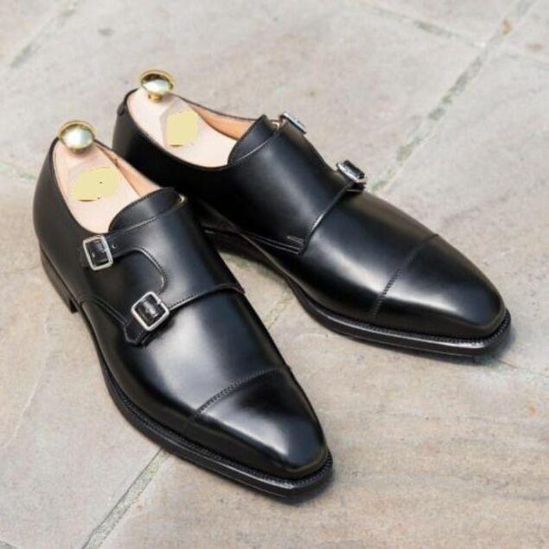 May include: A pair of black leather dress shoes with a double monk strap design. The shoes have a polished finish, a cap toe, and silver-colored buckles. Shoe trees are inserted inside the shoes.