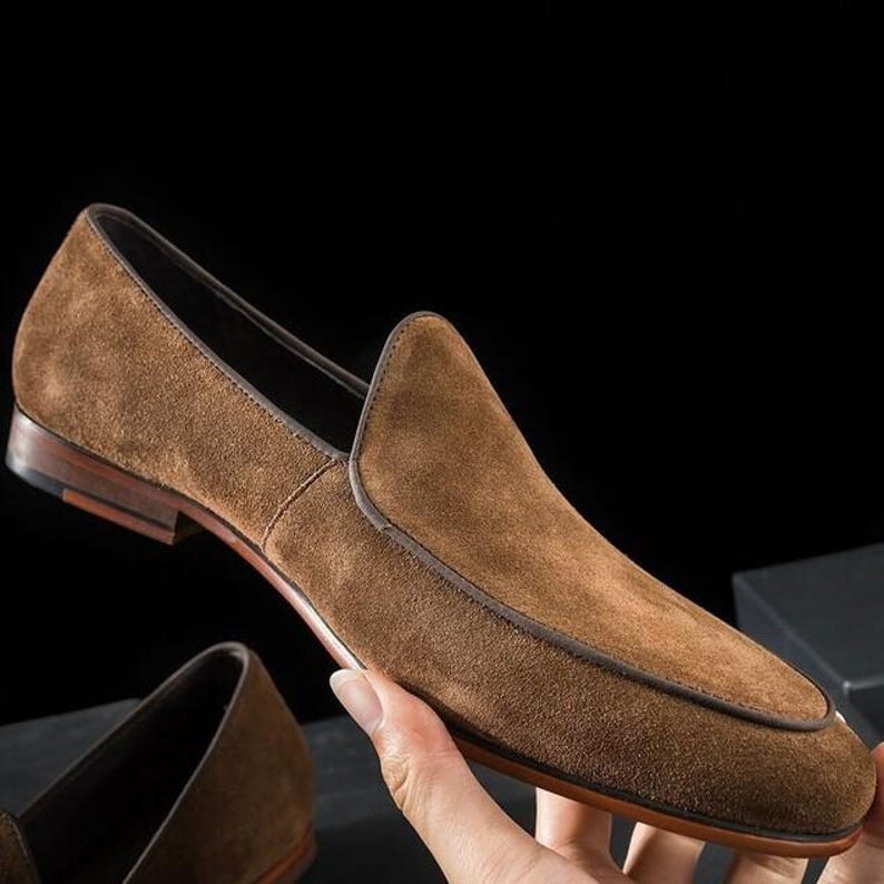 Puede incluir: Primer plano de un mocas&iacute;n de ante marr&oacute;n con punta afilada. El zapato tiene suela de cuero y ribete marr&oacute;n oscuro. El zapato est&aacute; sostenido por una mano sobre un fondo negro.