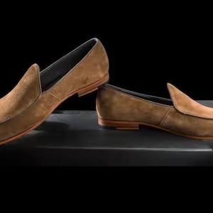 Puede incluir: Un par de mocasines de ante marr&oacute;n. Los zapatos tienen un dise&ntilde;o cl&aacute;sico sin cordones con punta redondeada y tac&oacute;n bajo. Las suelas son de color marr&oacute;n claro y los zapatos est&aacute;n sobre un fondo oscuro.