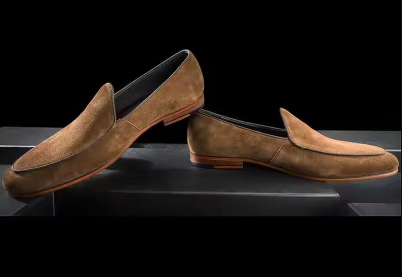 Puede incluir: Un par de mocasines de ante marr&oacute;n. Los zapatos tienen un dise&ntilde;o cl&aacute;sico sin cordones con punta redondeada y tac&oacute;n bajo. Las suelas son de color marr&oacute;n claro y los zapatos est&aacute;n sobre un fondo oscuro.