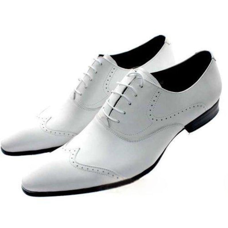 May include: A pair of white leather dress shoes with a classic wingtip design. The shoes feature a lace-up closure, a pointed toe, and black soles. The shoes are designed for formal occasions.