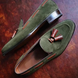 May include: A pair of olive green suede loafers with brown leather accents and tassels. The shoes have a classic slip-on design with a rounded toe and a low heel. The stitching and detailing are visible.