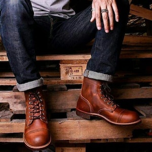 May include: Brown leather boots with laces and a rounded toe. The boots have a classic design and are worn with dark blue denim jeans. The boots are sitting on a wooden pallet.