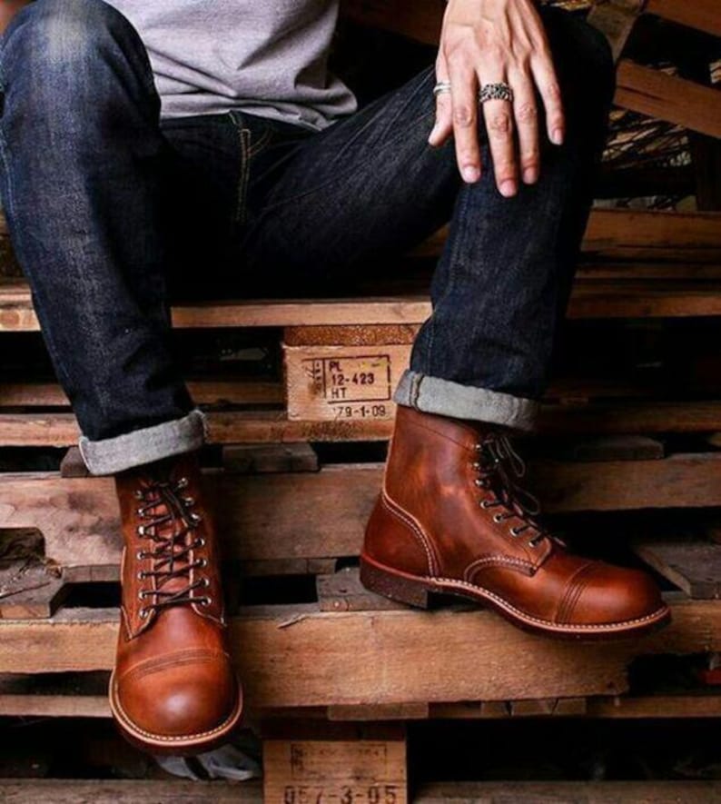 May include: Brown leather boots with laces and a rounded toe. The boots have a classic design and are worn with dark blue denim jeans. The boots are sitting on a wooden pallet.