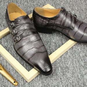 K&ouml;nnte beinhalten: Ein Paar graue Lederschuhe mit Krokodilmuster und drei Schnallenverschl&uuml;ssen. Die Schuhe werden auf einem Holzrahmen mit einem goldenen Schuhanzieher pr&auml;sentiert.
