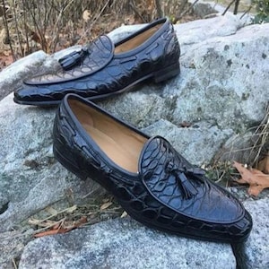 May include: Two black leather loafers with a textured, crocodile-effect pattern. Each shoe has a decorative tassel and a rounded toe. The shoes are presented on a grey stone surface, highlighting their design and craftsmanship.