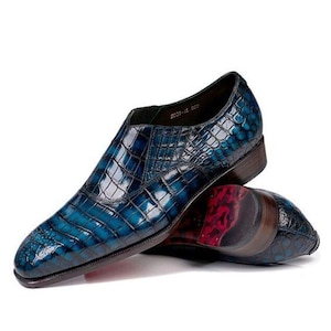 May include: A pair of blue leather dress shoes with a crocodile skin pattern. The shoes have a glossy finish and a dark brown sole. The sole of the shoe has a red and black pattern. The shoes are on a white background.