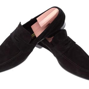 May include: A pair of black suede loafers with a slip-on design. The shoes feature a rounded toe and a strap across the top. A wooden shoe tree is inserted in one of the shoes. The shoes are on a white background.