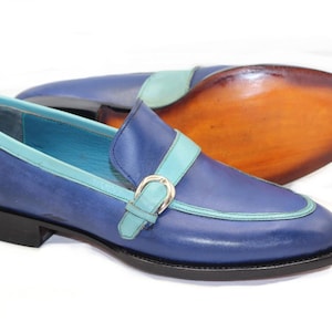 May include: Blue leather loafers with a light blue accent on the trim and strap. The shoes have a silver buckle and a black sole. The sole of the other shoe is visible, showing a brown leather sole.