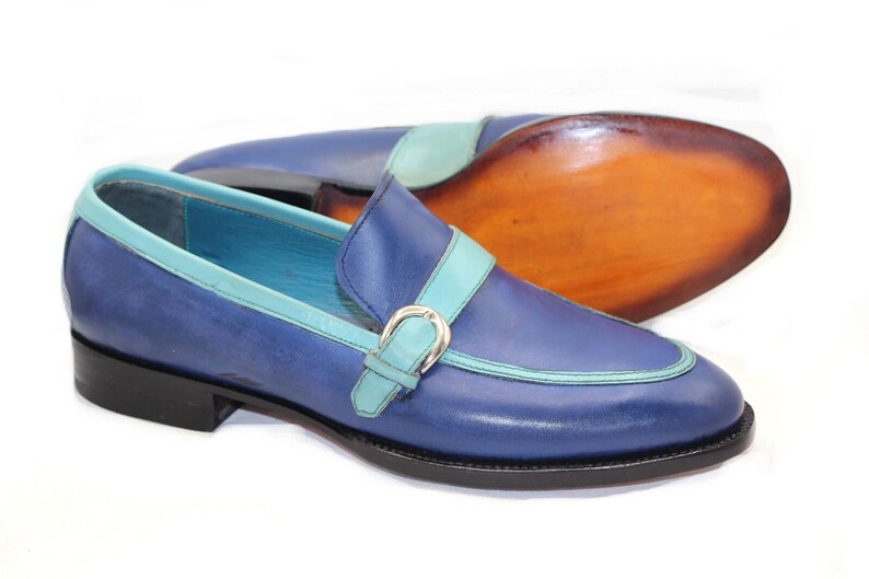 May include: Blue leather loafers with a light blue accent on the trim and strap. The shoes have a silver buckle and a black sole. The sole of the other shoe is visible, showing a brown leather sole.