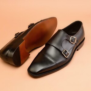May include: A pair of black leather double monk strap shoes with gold-colored buckles. The shoes have a square toe design and a brown sole. One shoe is positioned to show the sole.