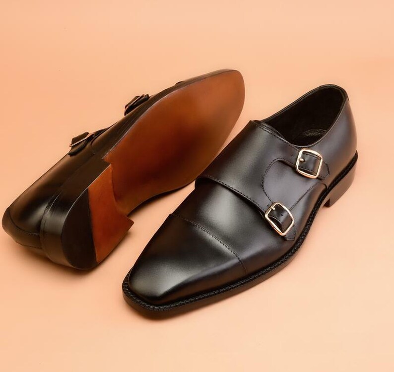 May include: A pair of black leather double monk strap shoes with gold-colored buckles. The shoes have a square toe design and a brown sole. One shoe is positioned to show the sole.