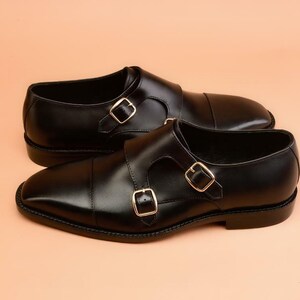 May include: A pair of black leather dress shoes with double monk straps and gold-colored buckles. The shoes have a polished finish and a classic design, suitable for formal occasions.