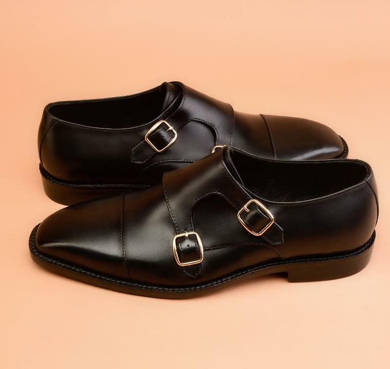 May include: A pair of black leather dress shoes with double monk straps and gold-colored buckles. The shoes have a polished finish and a classic design, suitable for formal occasions.