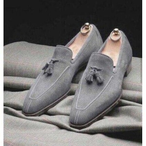 Puede incluir: Un par de mocasines grises de ante con detalles de borlas. Los zapatos tienen un dise&ntilde;o cl&aacute;sico con punta afilada y costuras blancas. Se insertan hormas. Los mocasines se muestran sobre tela gris.