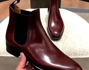 May include: A pair of burgundy Chelsea boots with a sleek, polished finish. The boots feature a pointed toe, elastic side panels, and a pull tab at the back. The sole is black with a thin white stripe. The boots are displayed on a white surface.