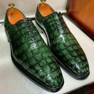 May include: A pair of green leather dress shoes with a textured, crocodile-like pattern. The shoes have a classic lace-up design, black soles, and a gold-colored shoe tree. The interior is a light brown color.