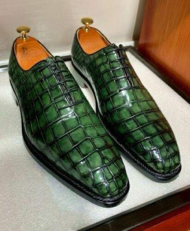 May include: A pair of green leather dress shoes with a textured, crocodile-like pattern. The shoes have a classic lace-up design, black soles, and a gold-colored shoe tree. The interior is a light brown color.