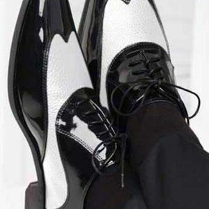 Può includere: Un paio di scarpe bicolore, nere e bianche, con finitura lucida. Le scarpe presentano un classico design a punta alata con chiusura con lacci. L'immagine mostra le scarpe da un'angolazione bassa, evidenziando il loro aspetto lucido.