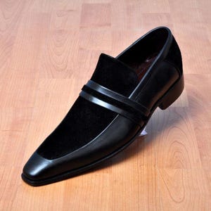 May include: Black leather slip-on shoe with a square toe and a velvet vamp. The shoe features two horizontal straps across the top and a low heel. The shoe is displayed on a wooden surface.
