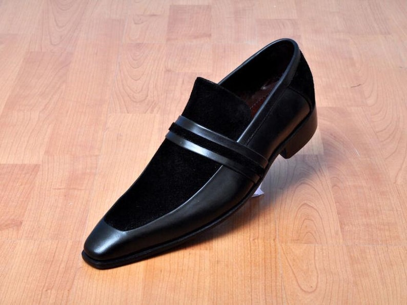 May include: Black leather slip-on shoe with a square toe and a velvet vamp. The shoe features two horizontal straps across the top and a low heel. The shoe is displayed on a wooden surface.