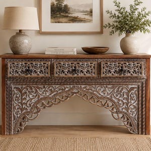 Può includere: Una consolle in legno con intricati disegni floreali intagliati. Il tavolo ha tre cassetti e una sezione inferiore ad arco. Una lampada, libri, una ciotola e un vaso con vegetazione sono sopra. La tavolozza dei colori comprende marrone, bianco e beige.