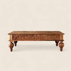 May include: A rectangular, brown wooden coffee table with ornate floral carvings along the front. The table has four rounded legs. The wood has a natural grain and a warm, inviting tone.