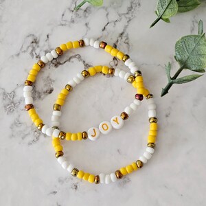 May include: Two beaded bracelets. One bracelet features the word "JOY" spelled out in white letter beads. The bracelets are made with white, yellow, and gold-colored beads. The bracelets are on a marble surface.