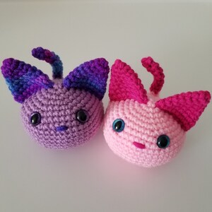 May include: Two crocheted cat plushies, one purple and blue with black eyes, the other pink with blue eyes.