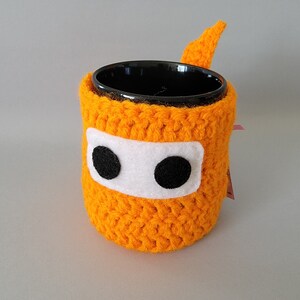 May include: Orange crocheted mug cozy with black eyes and a white stripe. The cozy is designed to fit a standard-sized mug.