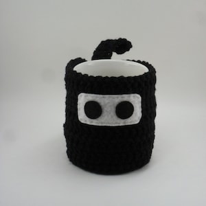 May include: Black crocheted mug cozy with a white felt face and two black felt eyes. The cozy has a long tail that wraps around the mug.