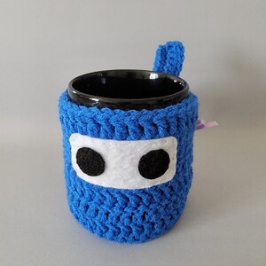 May include: A blue crocheted mug cozy with a white felt face and black felt eyes. The cozy is on a black mug.