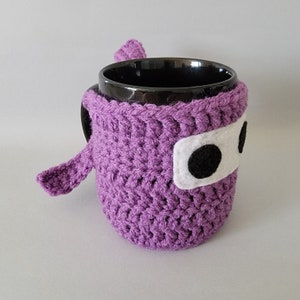 May include: Purple crocheted mug cozy with a white felt face and black felt eyes. The cozy is shaped like a ninja and has a long arm that wraps around the mug.