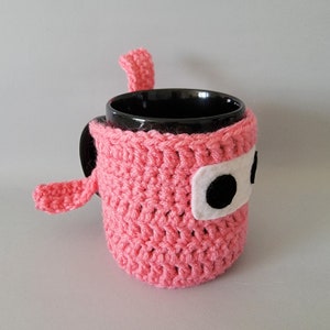 May include: A pink crocheted mug cozy with a white felt face and black felt eyes. The cozy is shaped like a cartoon character with a long, pointed nose.