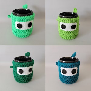 May include: Four crocheted mug cozies in green and blue. Each cozy has a black and white eye design. The cozies are made with a textured stitch.