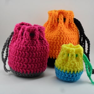 May include: Three crocheted drawstring bags in different sizes. The largest bag is orange, the middle bag is pink with a grey bottom, and the smallest bag is yellow with a blue bottom. The bags have black, grey, and green drawstrings.