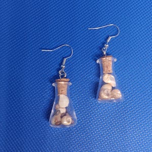 May include: A pair of unique earrings featuring small glass vials filled with seashells. Each vial is sealed with a cork stopper and suspended from silver-colored fish hook ear wires. The earrings are set against a blue background.