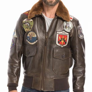 May include: Brown leather bomber jacket with a fur collar and multiple patches. The jacket has a zip closure, two front pockets, and ribbed cuffs and hem. Patches feature emblems and text, including "U.S.S. WILLIAM H. STANDLEY". The jacket's style is reminiscent of a classic flight jacket.