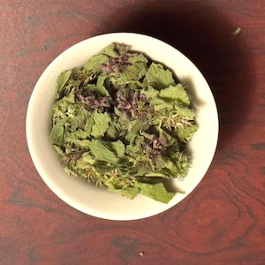 Könnte beinhalten: Eine kleine, weiße Keramikschale gefüllt mit getrockneten grünen Blättern und lila Blütenknospen. Die Schale befindet sich auf einer dunkelrotbraunen Holzoberfläche, was eine natürliche Ästhetik erzeugt. Die getrockneten Kräuter scheinen eine Teemischung zu sein.