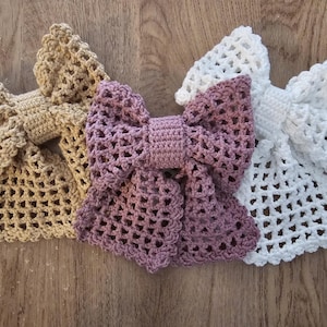 Könnte beinhalten: Vier handgefertigte Häkel-Schleifen in Beige, Mauve und Weiß. Die Schleifen haben ein offenes Webmuster. Sie liegen auf einer Holzoberfläche, was ihre Textur und Handwerkskunst hervorhebt. Jede Schleife ist ein einzigartiges Accessoire.
