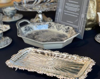 May include: A rectangular silver-colored tray with a starburst design in the center and a decorative, beaded edge. The tray has small feet and is displayed on a black surface. Other serving dishes are in the background.