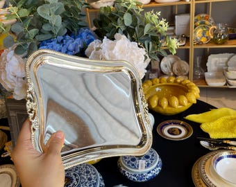 May include: A silver-colored, square serving tray with ornate detailing on the corners and edges. The tray is held in front of a display of decorative porcelain dishes, blue and white ceramic jars, and floral arrangements.