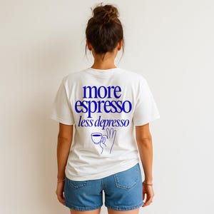 Könnte beinhalten: Weißes T-Shirt mit dem blauen Text "more espresso less depresso" und einer blauen Illustration einer Hand, die eine Kaffeetasse hält. Das Shirt wird mit Jeansshorts getragen.