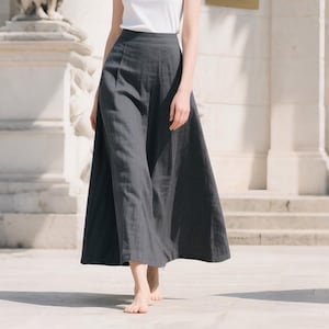 Puede incluir: Una falda de pierna ancha gris carbón hecha de una tela fluida. La falda se combina con una parte superior blanca. La falda tiene una cintura alta y llega hasta los tobillos. La modelo está descalza y camina sobre una superficie de piedra.