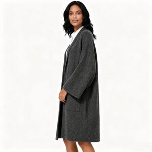 May include: A long, charcoal gray coat with wide sleeves and two front pockets. The coat is worn over a white collared shirt. The woman is standing in front of a white background.