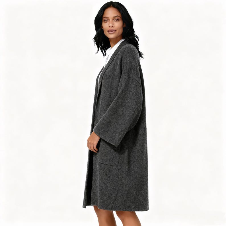 May include: A long, charcoal gray coat with wide sleeves and two front pockets. The coat is worn over a white collared shirt. The woman is standing in front of a white background.