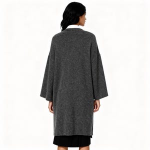 May include: A long, gray, oversized coat with wide sleeves. The coat is made of a soft, textured fabric and is worn over a black skirt and a white collared shirt. The model is facing away from the camera.