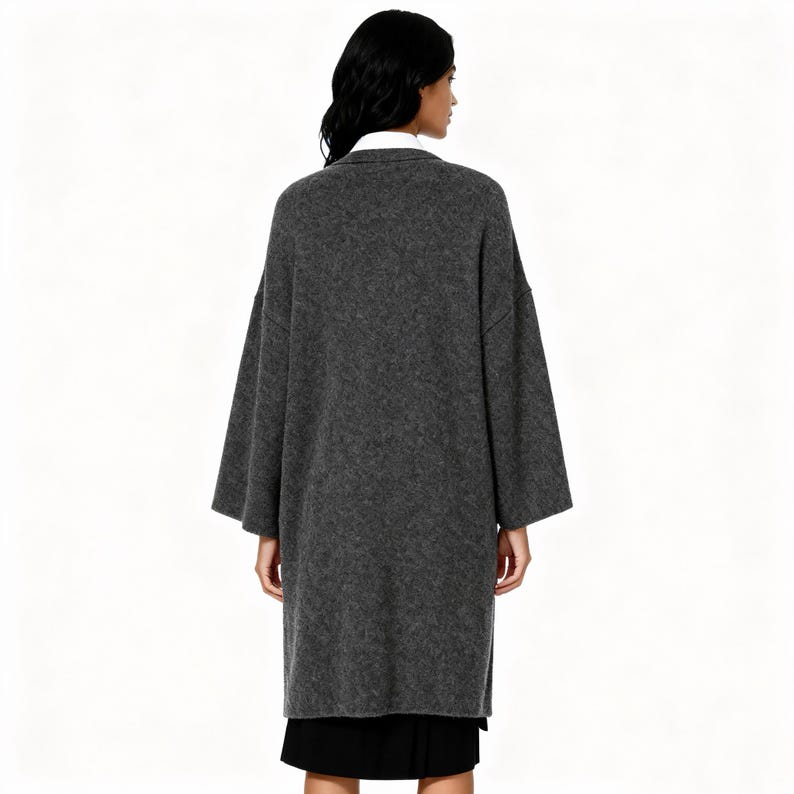 May include: A long, gray, oversized coat with wide sleeves. The coat is made of a soft, textured fabric and is worn over a black skirt and a white collared shirt. The model is facing away from the camera.